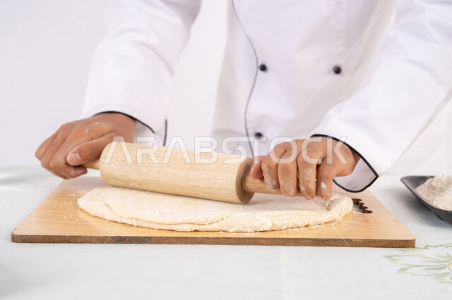 A Saudi chef preparing ingredients for making bread and breakfast, preparing and baking delicious baked goods and pastries, a close-up portrait of an Emirati Gulf Arab man wearing work clothes rolling out dough using a rolling pin, white background