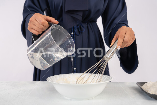 طهي المخبوزات والمعجنات الشهية، بورتريه مقرب لامرأة عربية خليجية اماراتية محجبة تقوم بخفق المكونات مع الدقيق الابيض لتحضير الطعام، ربة منزل سعودية تقوم بتجهيز المكونات لصناعة الخبز والفطور، خلفية بيضاء