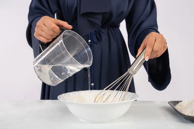 طهي المخبوزات والمعجنات الشهية، بورتريه مقرب لامرأة عربية خليجية اماراتية محجبة تقوم بخفق المكونات مع الدقيق الابيض لتحضير الطعام، ربة منزل سعودية تقوم بتجهيز المكونات لصناعة الخبز والفطور، خلفية بيضاء