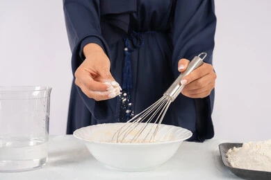 Preparing delicious baked goods and pastries, using a rolling pin. Close-up portrait of a veiled Emirati Gulf Arab woman mixing ingredients with white flour to prepare food. A Saudi housewife preparing ingredients for making bread and breakfast, white background.