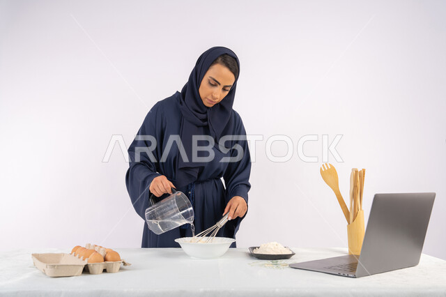 مشاركة الوصفة عبر المواقع باستخدام الحاسوب المحمول، إعداد المخبوزات والمعجنات الشهية، بورتريه مقرب لامرأة عربية خليجية اماراتية محجبة تقوم بخفق المكونات مع الدقيق الابيض لتحضير الطعام، ربة منزل سعودية تقوم بتجهيز المكونات لصناعة الخبز والفطور، خلفية بيضاء