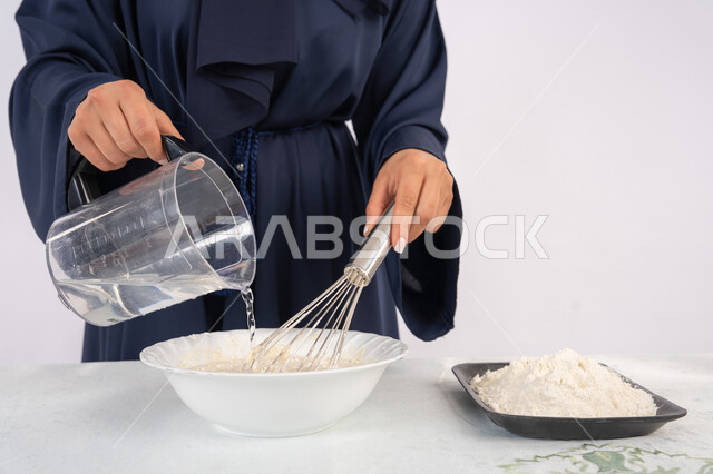 إعداد المخبوزات والمعجنات الشهية، بورتريه مقرب لامرأة عربية خليجية اماراتية محجبة تقوم بخفق المكونات مع الدقيق الابيض لتحضير الطعام، ربة منزل سعودية تقوم بتجهيز المكونات لصناعة الخبز والفطور، خلفية بيضاء