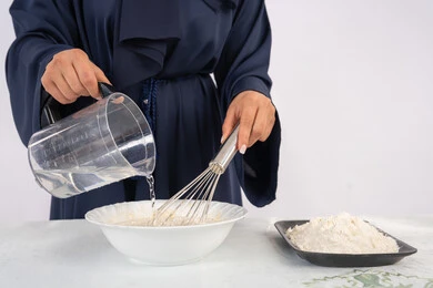 إعداد المخبوزات والمعجنات الشهية، بورتريه مقرب لامرأة عربية خليجية اماراتية محجبة تقوم بخفق المكونات مع الدقيق الابيض لتحضير الطعام، ربة منزل سعودية تقوم بتجهيز المكونات لصناعة الخبز والفطور، خلفية بيضاء