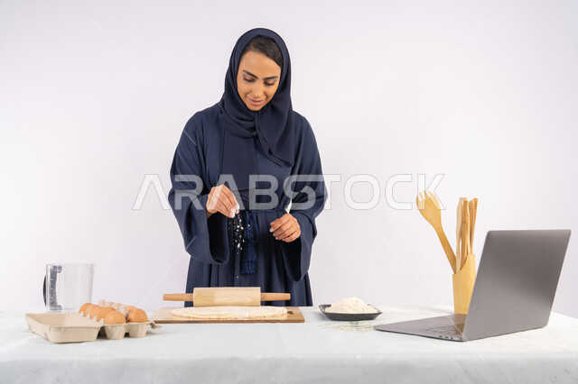 تحضير وإعداد المخبوزات والمعجنات الشهية، مشاركة الوصفة عبر المواقع باستخدام الحاسوب المحمول، بورتريه مقرب لامرأة عربية خليجية اماراتية محجبة تقوم تقوم بفرد العجين باستخدام الشوبك (النشابة)، ربة منزل سعودية تقوم بتجهيز المكونات لصناعة الخبز والفطور، خلفية بيضاء