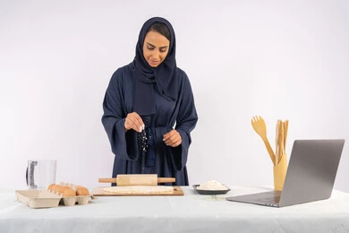 تحضير وإعداد المخبوزات والمعجنات الشهية، مشاركة الوصفة عبر المواقع باستخدام الحاسوب المحمول، بورتريه مقرب لامرأة عربية خليجية اماراتية محجبة تقوم تقوم بفرد العجين باستخدام الشوبك (النشابة)، ربة منزل سعودية تقوم بتجهيز المكونات لصناعة الخبز والفطور، خلفية بيضاء