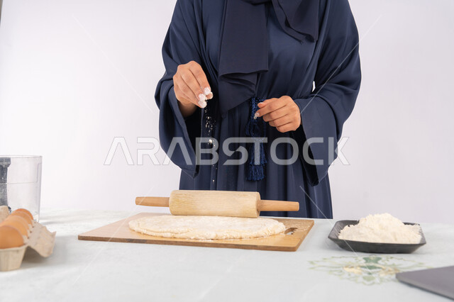 A Saudi housewife preparing ingredients for making bread and breakfast, preparing and baking delicious baked goods and pastries, a close-up portrait of a veiled Emirati Gulf Arab woman rolling out dough using a rolling pin, white background