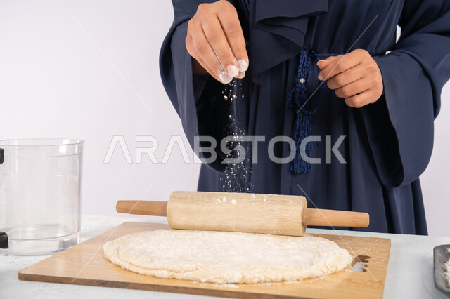 شيف سعودية تقوم بتجهيز المكونات لصناعة الخبز والفطور، تحضير وإعداد المخبوزات والمعجنات الشهية، بورتريه مقرب لامرأة عربية خليجية اماراتية محجبة تقوم تقوم بفرد العجين باستخدام الشوبك (النشابة)، خلفية بيضاء