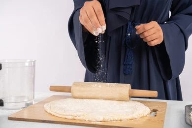 A Saudi housewife preparing ingredients for making bread and breakfast, preparing and baking delicious baked goods and pastries, a close-up portrait of a veiled Emirati Gulf Arab woman rolling out dough using a rolling pin, white background