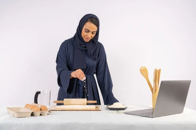 A Saudi housewife preparing ingredients for making bread and breakfast, preparing and baking delicious baked goods and pastries, a close-up portrait of a veiled Emirati Gulf Arab woman rolling out dough using a rolling pin, white background