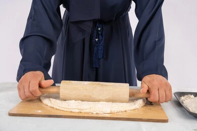 طباخة سعودية تقوم بتجهيز المكونات لصناعة الخبز والفطور، تحضير وإعداد المخبوزات والمعجنات الشهية، بورتريه مقرب لامرأة عربية خليجية اماراتية محجبة تقوم بفرد العجين باستخدام الشوبك (النشابة)، خلفية بيضاء