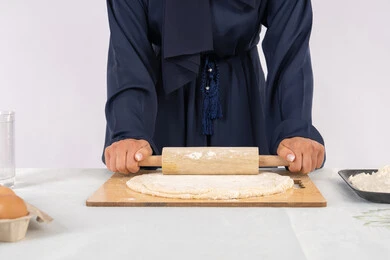 A Saudi housewife preparing ingredients for making bread and breakfast, preparing and baking delicious baked goods and pastries, a close-up portrait of a veiled Emirati Gulf Arab woman rolling out dough using a rolling pin, white background