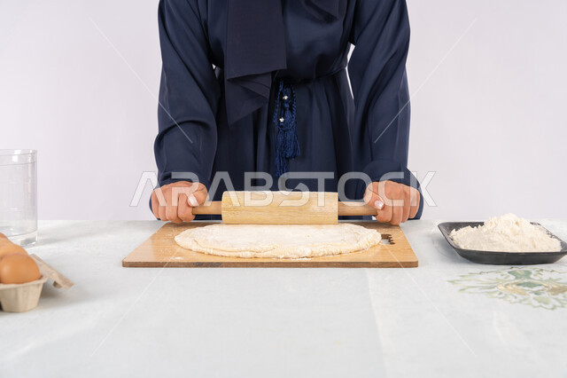 طباخة سعودية تقوم بتجهيز المكونات لصناعة الخبز والفطور، تحضير وإعداد المخبوزات والمعجنات الشهية، بورتريه مقرب لامرأة عربية خليجية اماراتية محجبة تقوم بفرد العجين باستخدام الشوبك (النشابة)، خلفية بيضاء