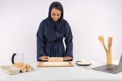 تحضير وإعداد المخبوزات والمعجنات الشهية، مشاركة الوصفة عبر المواقع باستخدام الحاسوب المحمول، بورتريه مقرب لامرأة عربية خليجية اماراتية محجبة تقوم تقوم بفرد العجين باستخدام الشوبك (النشابة)، ربة منزل سعودية تقوم بتجهيز المكونات لصناعة الخبز والفطور، خلفية بيضاء