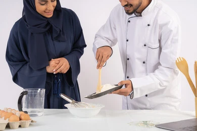التدرب على المقادير والمكونات المعيارية، ربة منزل سعودية تقوم بتعلم صناعة الخبز والمعجنات،  دورة تحضير وإعداد المخبوزات الشهية، بورتريه لشيف عربي خليجي اماراتي يقوم بتدريب إمرأة على اساسيات العمل في المطبخ، خلفية بيضاء