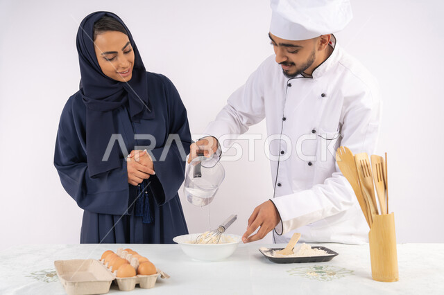 التدرب على المقادير والمكونات المعيارية، ربة منزل سعودية تقوم بتعلم صناعة الخبز والمعجنات،  دورة تحضير وإعداد المخبوزات الشهية، بورتريه لشيف عربي خليجي اماراتي يقوم بتدريب إمرأة على اساسيات العمل في المطبخ، خلفية بيضاء