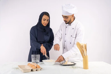 التدرب على المقادير والمكونات المعيارية، ربة منزل سعودية تقوم بتعلم صناعة الخبز والمعجنات،  دورة تحضير وإعداد المخبوزات الشهية، بورتريه لشيف عربي خليجي اماراتي يقوم بتدريب إمرأة على اساسيات العمل في المطبخ، خلفية بيضاء