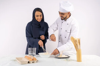 التدرب على المقادير والمكونات المعيارية، ربة منزل سعودية تقوم بتعلم صناعة الخبز والمعجنات،  دورة تحضير وإعداد المخبوزات الشهية، بورتريه لشيف عربي خليجي اماراتي يقوم بتدريب إمرأة على اساسيات العمل في المطبخ، خلفية بيضاء