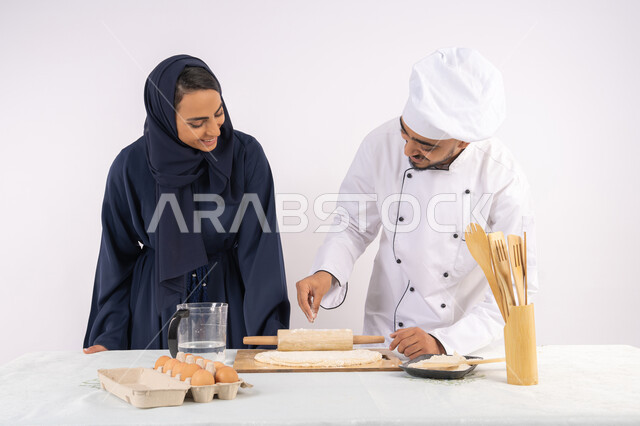 اساسيات العمل في المطبخ، ربة منزل سعودية تقوم بتعلم صناعة الخبز والمعجنات، دورة تحضير وإعداد المخبوزات الشهية، بورتريه لشيف عربي خليجي اماراتي يقوم بتدريب إمرأة على مهارة فرد العجين باستخدام الشوبك (النشابة) ، خلفية بيضاء