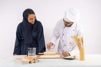 اساسيات العمل في المطبخ، ربة منزل سعودية تقوم بتعلم صناعة الخبز والمعجنات، دورة تحضير وإعداد المخبوزات الشهية، بورتريه لشيف عربي خليجي اماراتي يقوم بتدريب إمرأة على مهارة فرد العجين باستخدام الشوبك (النشابة) ، خلفية بيضاء