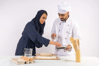 اساسيات العمل في المطبخ، ربة منزل سعودية تقوم بتعلم صناعة الخبز والمعجنات، دورة تحضير وإعداد المخبوزات الشهية، بورتريه لشيف عربي خليجي اماراتي يقوم بتدريب إمرأة على مهارة فرد العجين باستخدام الشوبك (النشابة) ، خلفية بيضاء