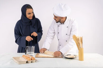 اساسيات العمل في المطبخ، ربة منزل سعودية تقوم بتعلم صناعة الخبز والمعجنات، دورة تحضير وإعداد المخبوزات الشهية، بورتريه لشيف عربي خليجي اماراتي يقوم بتدريب إمرأة على مهارة فرد العجين باستخدام الشوبك (النشابة) ، خلفية بيضاء