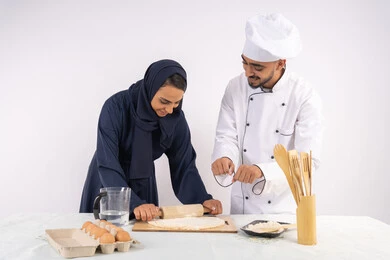 طباخة سعودية تقوم بتعلم صناعة الخبز والمعجنات،  دورة تحضير وإعداد المخبوزات الشهية، بورتريه لشيف عربي خليجي اماراتي يقوم بتدريب إمرأة على اساسيات العمل في المطبخ، مهارة فرد العجين باستخدام الشوبك (النشابة)، خلفية بيضاء