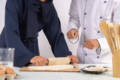 اساسيات العمل في المطبخ، ربة منزل سعودية تقوم بتعلم صناعة الخبز والمعجنات، دورة تحضير وإعداد المخبوزات الشهية، بورتريه لشيف عربي خليجي اماراتي يقوم بتدريب إمرأة على مهارة فرد العجين باستخدام الشوبك (النشابة) ، خلفية بيضاء