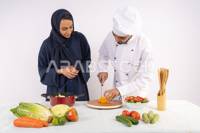 فنون تقطيع الخضراوات، ربة منزل سعودية تقوم بتعلم الطهي وإعداد الأطباق، دورة تحضير وصفات السلطات والوجبات الصحية الشهية، بورتريه لشيف عربي خليجي اماراتي يقوم بتدريب إمرأة على اساسيات العمل في المطبخ، خلفية بيضاء