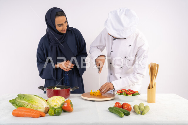 احترافية تقطيع الخضراوات، ربة منزل سعودية تقوم بتعلم الطهي وإعداد الأطباق، دورة تحضير وصفات السلطات والوجبات الصحية الشهية، بورتريه لشيف عربي خليجي اماراتي يقوم بتدريب إمرأة على اساسيات العمل في المطبخ، خلفية بيضاء