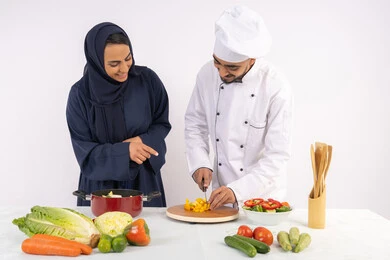 تحضير وصفات السلطات والوجبات الصحية الشهية، بورتريه لشيف عربي خليجي اماراتي يقوم بتدريب إمرأة على اساسيات العمل في المطبخ، تعلم مهارة تقطيع الخضراوات، ربة منزل سعودية تقوم بتعلم الطهي وإعداد الأطباق، خلفية بيضاء
