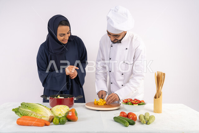 اساليب تقطيع الخضراوات، ربة منزل سعودية تقوم بتعلم الطهي وإعداد الأطباق، دورة تحضير وصفات السلطات والوجبات الصحية الشهية، بورتريه لشيف عربي خليجي اماراتي يقوم بتدريب إمرأة على اساسيات العمل في المطبخ، خلفية بيضاء