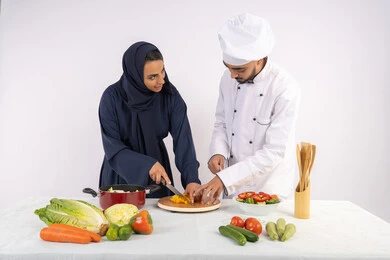 A Saudi cook is learning to cook and prepare dishes. A course is being held to prepare recipes for salads and delicious healthy meals. A portrait of an Emirati Gulf Arab chef is training a woman on the basics of working in the kitchen. She is learning the skill of cutting vegetables. White background.