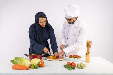 طباخة سعودية تقوم بتعلم الطهي وإعداد الأطباق، دورة تحضير وصفات السلطات والوجبات الصحية الشهية، بورتريه لشيف عربي خليجي اماراتي يقوم بتدريب إمرأة على اساسيات العمل في المطبخ، تعلم مهارة تقطيع الخضراوات،  خلفية بيضاء