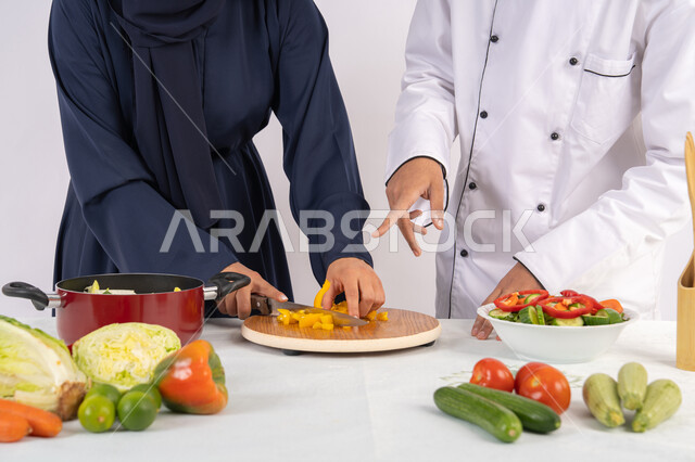 دورة تحضير وصفات السلطات والوجبات الصحية الشهية، بورتريه مقرب لشيف عربي خليجي اماراتي يقوم بتدريب إمرأة على اساسيات العمل في المطبخ، تعلم مهارة تقطيع الخضراوات، ربة منزل سعودية تقوم بتعلم الطهي وإعداد الأطباق، خلفية بيضاء