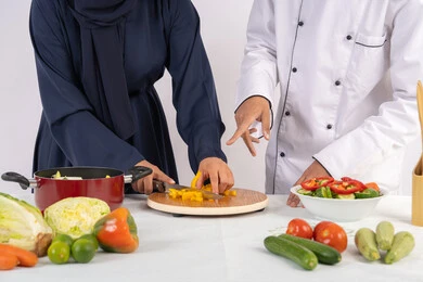 دورة تحضير وصفات السلطات والوجبات الصحية الشهية، بورتريه مقرب لشيف عربي خليجي اماراتي يقوم بتدريب إمرأة على اساسيات العمل في المطبخ، تعلم مهارة تقطيع الخضراوات، ربة منزل سعودية تقوم بتعلم الطهي وإعداد الأطباق، خلفية بيضاء