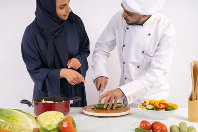دورة تحضير وصفات السلطات والوجبات الصحية الشهية، بورتريه لشيف عربي خليجي اماراتي يقوم بتدريب إمرأة على اساسيات العمل في المطبخ، تعلم مهارة تقطيع الخضراوات، ربة منزل سعودية تقوم بتعلم الطهي وإعداد الأطباق، خلفية بيضاء