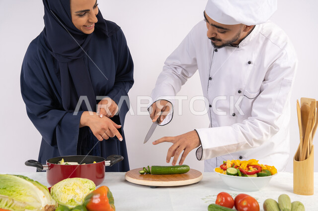 تعلم مهارة تقطيع الخضراوات، ربة منزل سعودية تقوم بتعلم الطهي وإعداد الأطباق، دورة تحضير وصفات السلطات والوجبات الصحية الشهية، بورتريه لشيف عربي خليجي اماراتي يقوم بتدريب إمرأة على اساسيات العمل في المطبخ، خلفية بيضاء