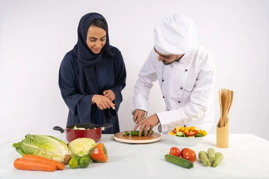 احترافية تقطيع الخضراوات، ربة منزل سعودية تقوم بتعلم الطهي وإعداد الأطباق، دورة تحضير وصفات السلطات والوجبات الصحية الشهية، بورتريه لشيف عربي خليجي اماراتي يقوم بتدريب إمرأة على اساسيات العمل في المطبخ، خلفية بيضاء