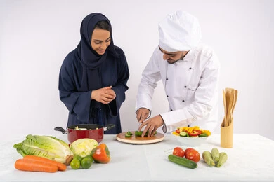 A course on preparing delicious, healthy salad and meal recipes. A portrait of an Emirati Gulf Arab chef training a woman on the basics of working in the kitchen. Learning the skill of cutting vegetables. A Saudi housewife learning to cook and prepare dishes. White background.