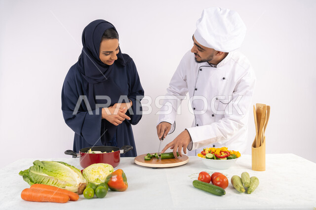 تعلم مهارة تقطيع الخضراوات، ربة منزل سعودية تقوم بتعلم الطهي وإعداد الأطباق، دورة تحضير وصفات السلطات والوجبات الصحية الشهية، بورتريه لشيف عربي خليجي اماراتي يقوم بتدريب إمرأة على اساسيات العمل في المطبخ، خلفية بيضاء