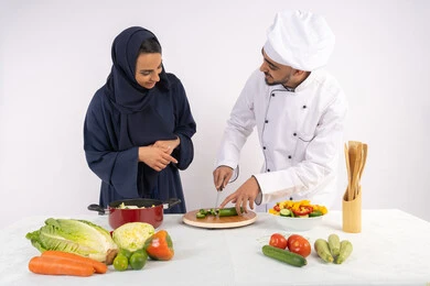 تعلم مهارة تقطيع الخضراوات، ربة منزل سعودية تقوم بتعلم الطهي وإعداد الأطباق، دورة تحضير وصفات السلطات والوجبات الصحية الشهية، بورتريه لشيف عربي خليجي اماراتي يقوم بتدريب إمرأة على اساسيات العمل في المطبخ، خلفية بيضاء