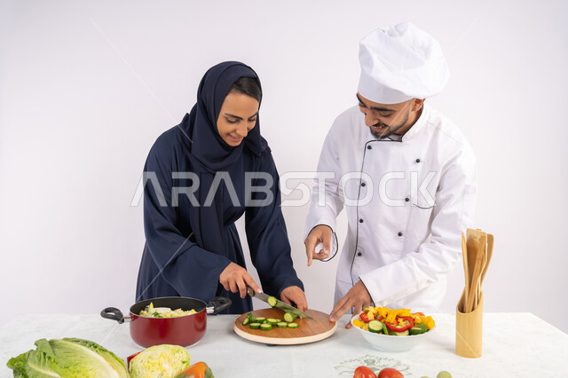 دورة تحضير وصفات السلطات والوجبات الصحية الشهية، بورتريه لشيف عربي خليجي اماراتي يقوم بتدريب إمرأة على اساسيات العمل في المطبخ، تعلم مهارة تقطيع الخضراوات، ربة منزل سعودية تقوم بتعلم الطهي وإعداد الأطباق، خلفية بيضاء