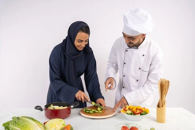 دورة تحضير وصفات السلطات والوجبات الصحية الشهية، بورتريه لشيف عربي خليجي اماراتي يقوم بتدريب إمرأة على اساسيات العمل في المطبخ، تعلم مهارة تقطيع الخضراوات، ربة منزل سعودية تقوم بتعلم الطهي وإعداد الأطباق، خلفية بيضاء