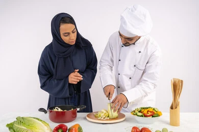 Learning the skill of cutting vegetables, a Saudi housewife learning to cook and prepare dishes, a course in preparing recipes for salads and delicious healthy meals, a portrait of an Arab Gulf Emirati chef training a woman on the basics of working in the kitchen, white background