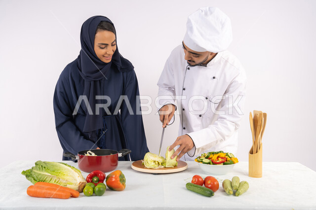 احترافية تقطيع الخضراوات، ربة منزل سعودية تقوم بتعلم الطهي وإعداد الأطباق، دورة تحضير وصفات السلطات والوجبات الصحية الشهية، بورتريه لشيف عربي خليجي اماراتي يقوم بتدريب إمرأة على اساسيات العمل في المطبخ، خلفية بيضاء
