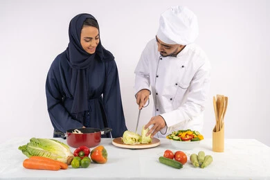 احترافية تقطيع الخضراوات، ربة منزل سعودية تقوم بتعلم الطهي وإعداد الأطباق، دورة تحضير وصفات السلطات والوجبات الصحية الشهية، بورتريه لشيف عربي خليجي اماراتي يقوم بتدريب إمرأة على اساسيات العمل في المطبخ، خلفية بيضاء