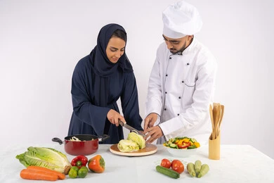 Learning the skill of cutting vegetables, a Saudi housewife learning to cook and prepare dishes, a course in preparing recipes for salads and delicious healthy meals, a portrait of an Arab Gulf Emirati chef training a woman on the basics of working in the kitchen, white background