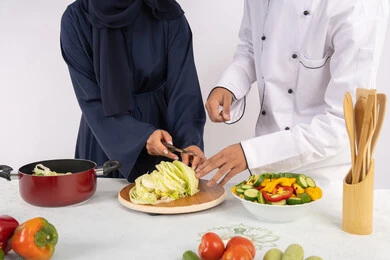 دورة تحضير وصفات السلطات والوجبات الصحية الشهية، بورتريه مقرب لشيف عربي خليجي اماراتي يقوم بتدريب إمرأة على اساسيات العمل في المطبخ، تعلم مهارة تقطيع الخضراوات، ربة منزل سعودية تقوم بتعلم الطهي وإعداد الأطباق، خلفية بيضاء