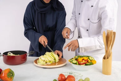 Learning the skill of cutting vegetables, a Saudi housewife learning to cook and prepare dishes, a course in preparing recipes for salads and delicious healthy meals, a portrait of an Arab Gulf Emirati chef training a woman on the basics of working in the kitchen, white background