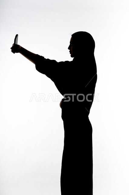 اجراء مكالمه هاتفيه ،التقاط صور سيلفي تذكاريه بإستخدام الهاتف المحمول ، صورة ظلية لمرأة عربية خليجية اماراتيه ، إستخدام اجهزة تقنية حديثة ، التواصل الإجتماعي ، بث مباشر ، خلفية بيضاء